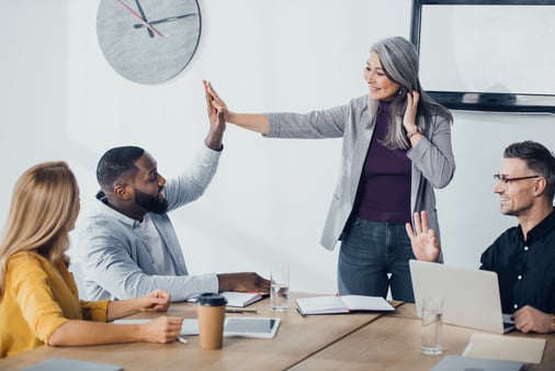 Four employees sitting or standing around a table. All are smiling while two of them are giving each other a high five.