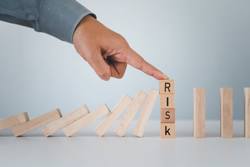 A person's finger holding in place a set of stacked blocks spelling out the word' risk' from falling in a line of dominos. 