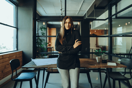 Confident business woman leaning against a desk in a modern office with her arms crossed, looking directly at the camera. 