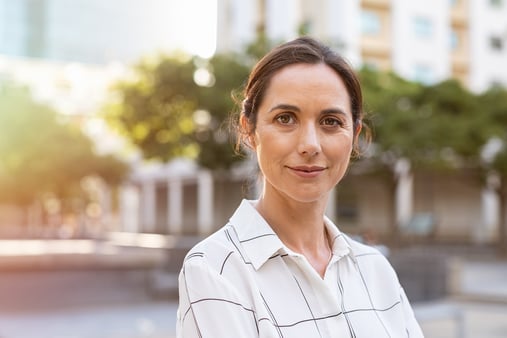 Confident, smiling business woman in front of an outdoor plaza with trees in the background.