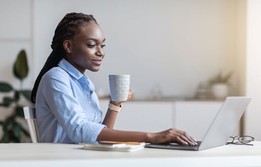 Woman with box braids and a blue shirt sitting at a desk drinking coffee and working on her computer 